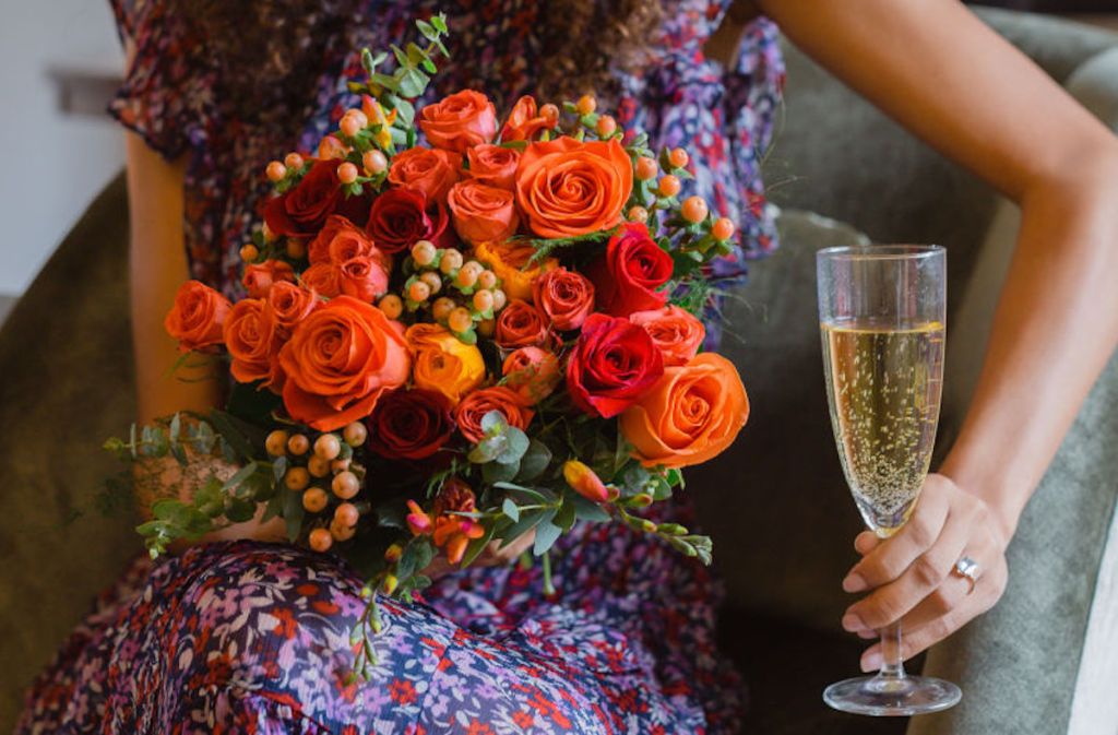 woman holding red and orange bouquet of flowers holding glass of champagne