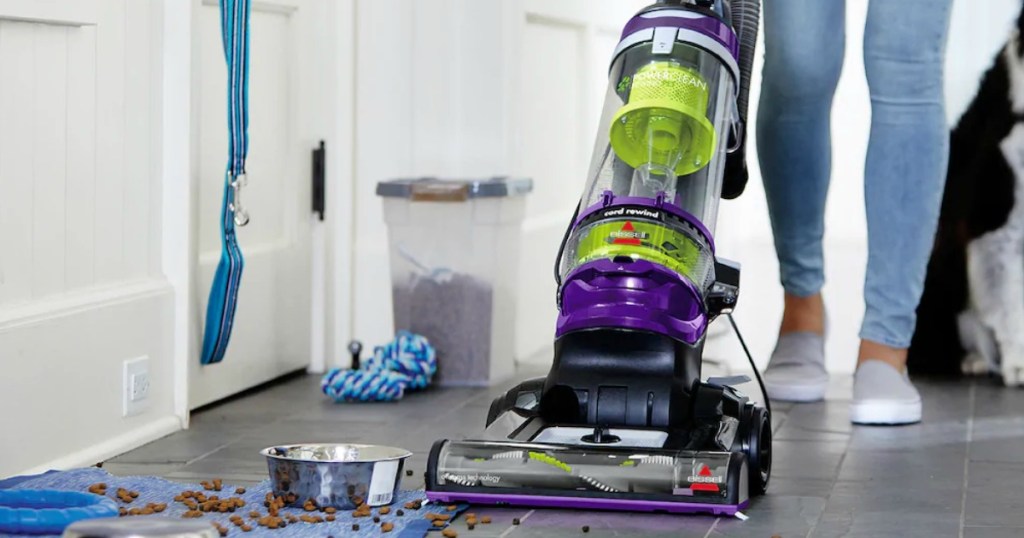 Woman using a large vacuum cleaner on hardwood floor near spilled dog food