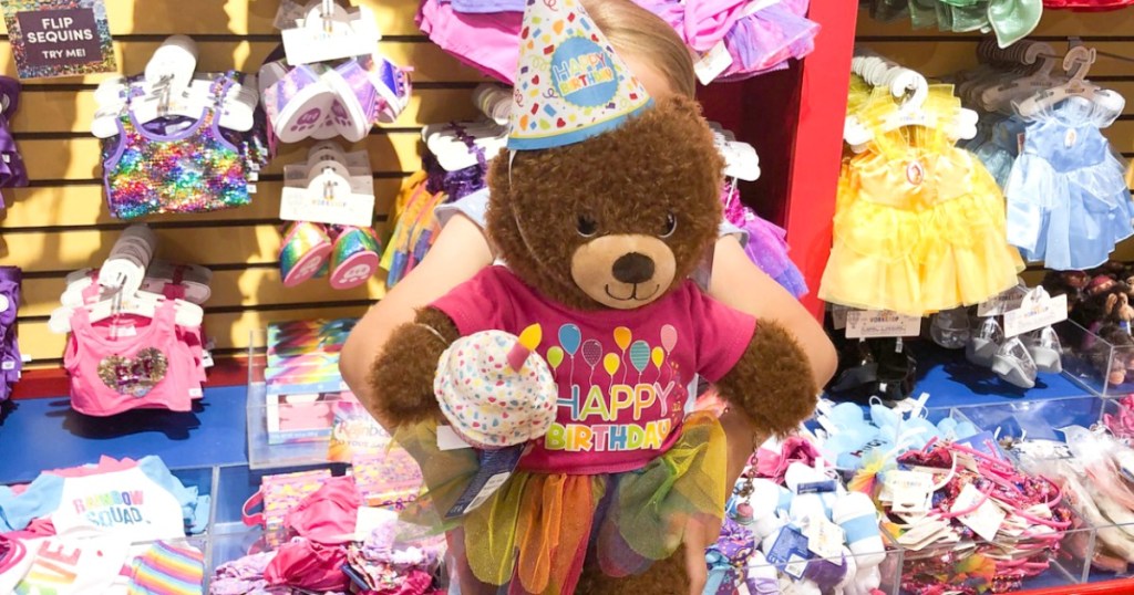 Young girl holding a large Birthday-themed teddy bear in front of her in a store