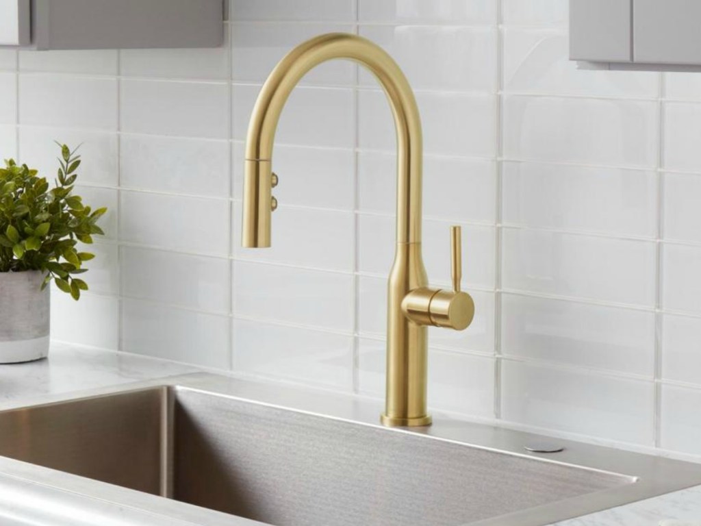 Large golden kitchen sink faucet on a sink near a subway tile backsplash and a small potted plant