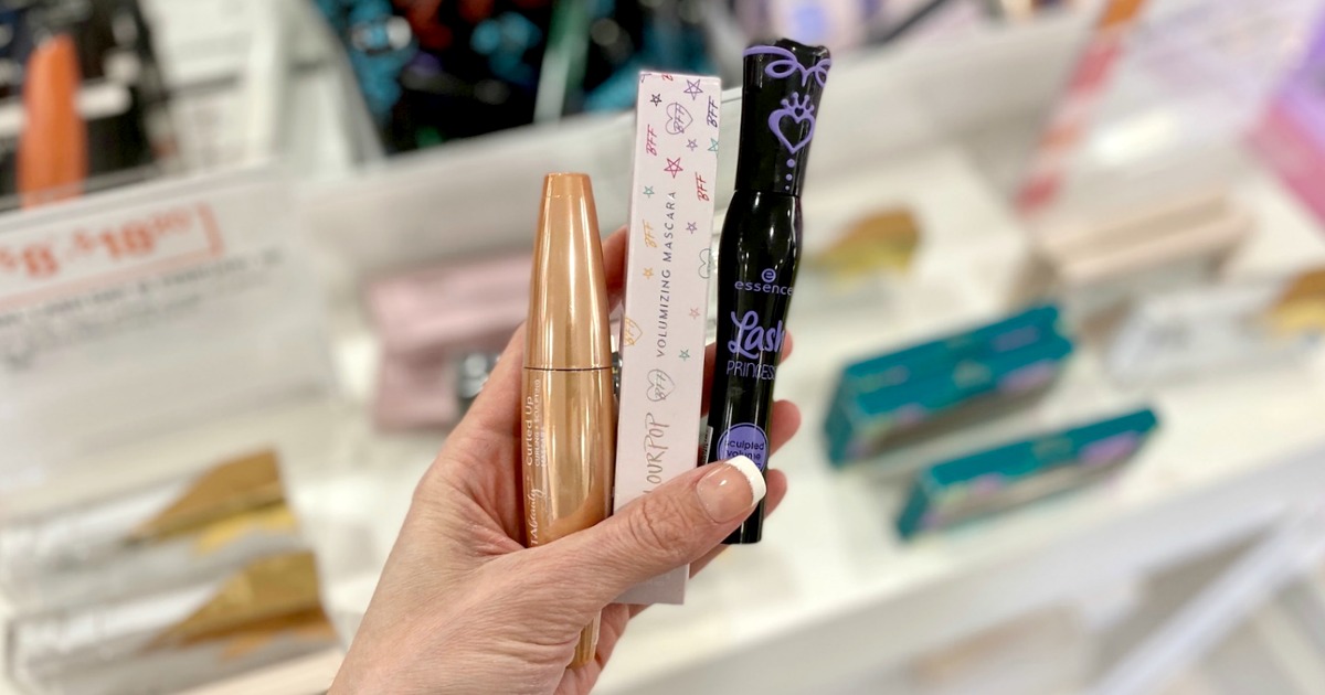 Hand holding three different mascaras in front of in-store display at beauty store