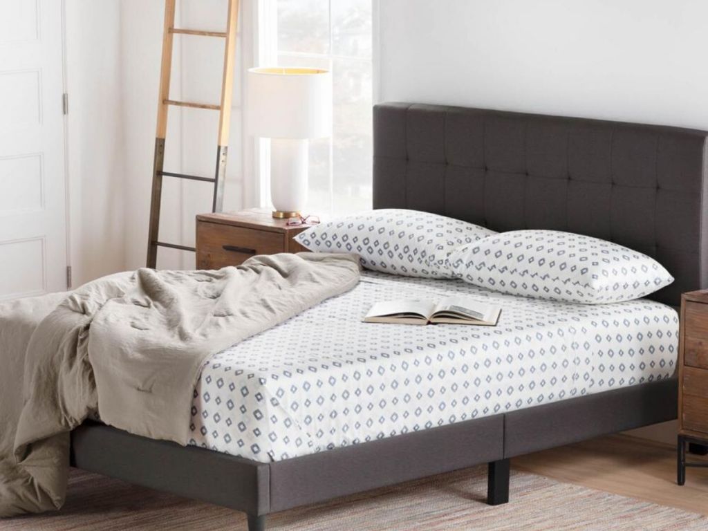 partially made bed with upholstered headboard with nightstand and decorative ladder on the left side of bed with window in the background