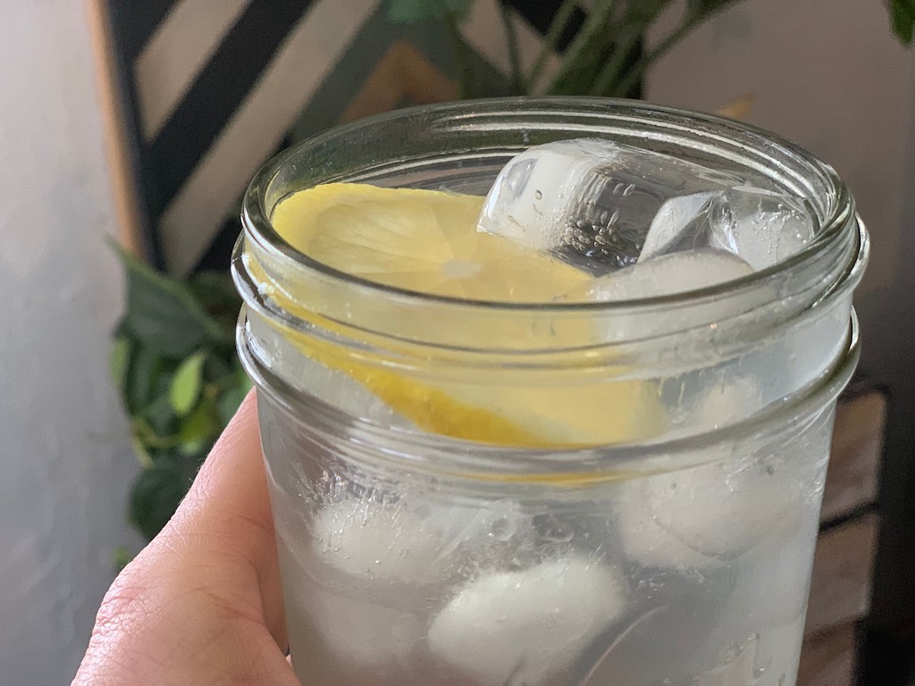 holding glass mason jar filled with water, ice, and lemon slice 