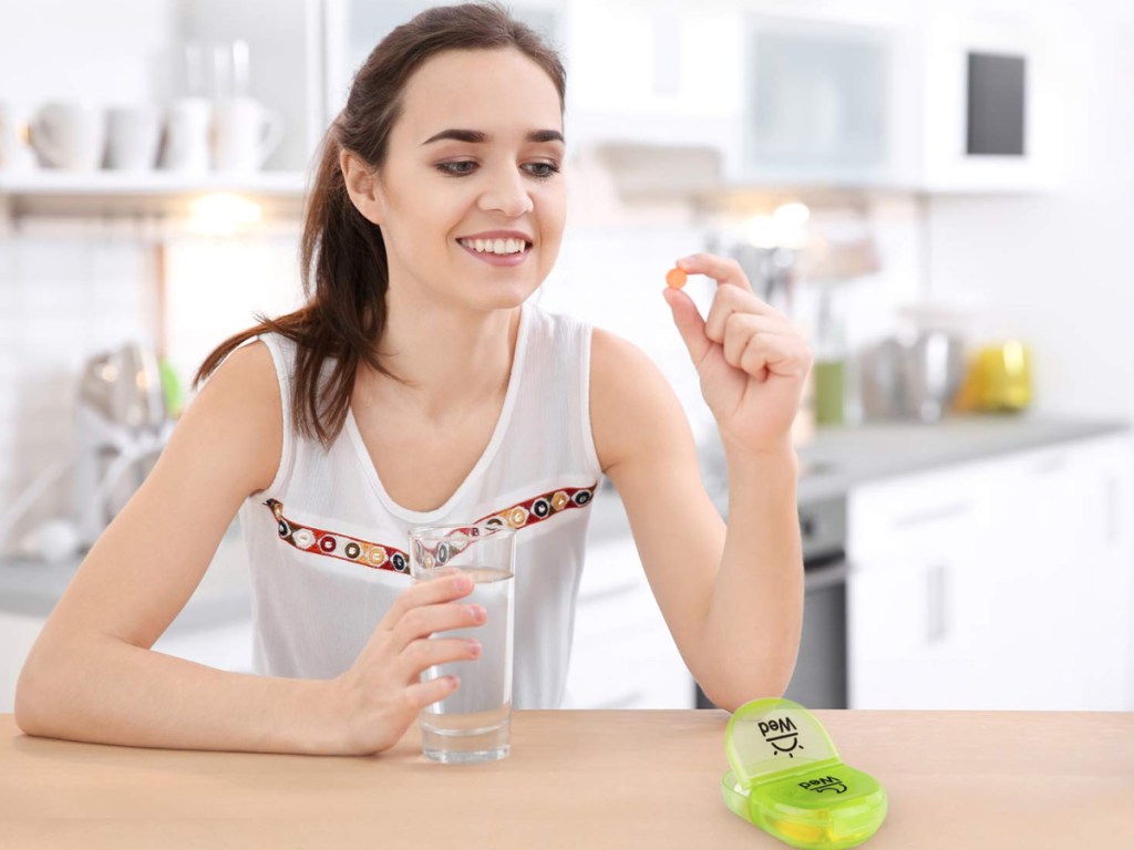 woman holding a glass of water in one hand and pill in other hand with green pill organizer on table