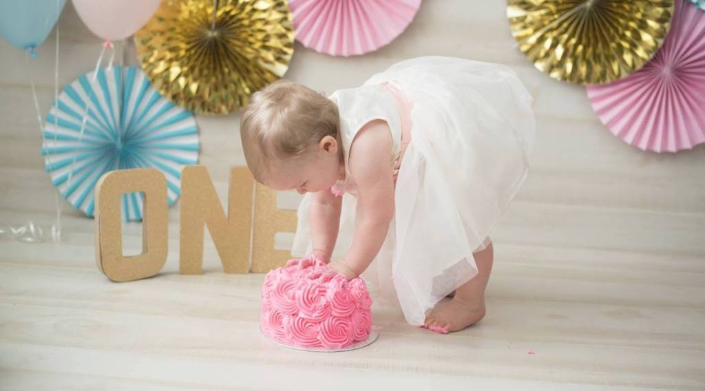baby girl wearing white dress touching rose pink icing on cake