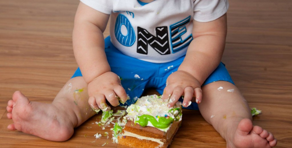 baby with one shirt sitting on wood floor touching piece of birthday cake 