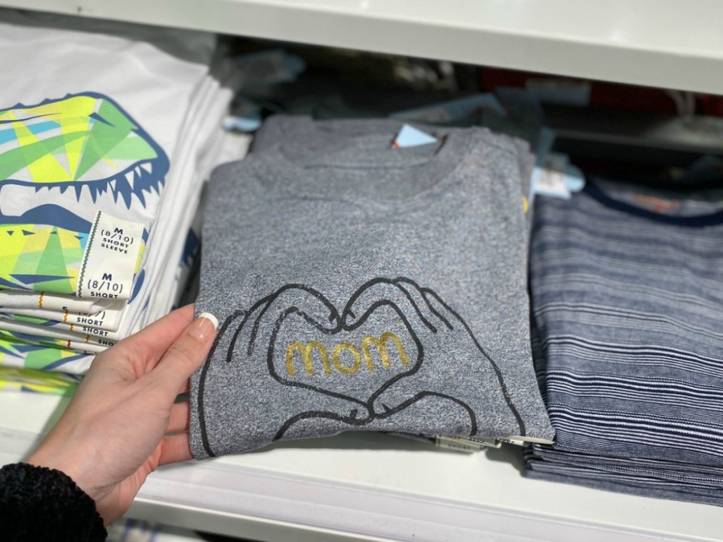 Woman holding a folded boys graphic tee that says "mom" near in-store display