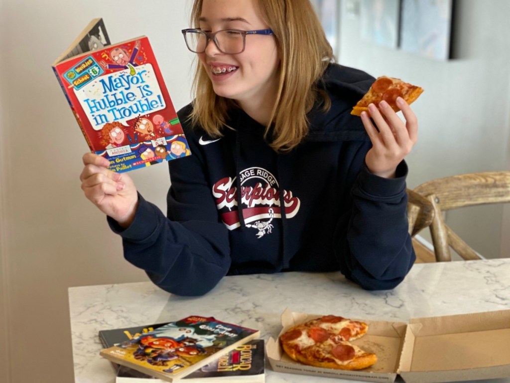 Preteen girl reading a book and holding a piece of pepperoni pizza