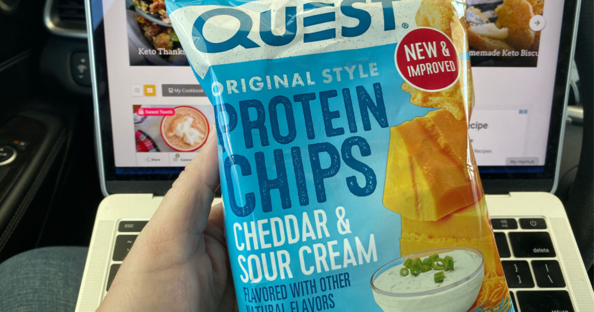 hand holding up quest protein chips cheddar and sour cream in front of laptop