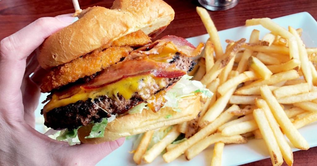 hand holding burger with bacon on it over a plate of fries