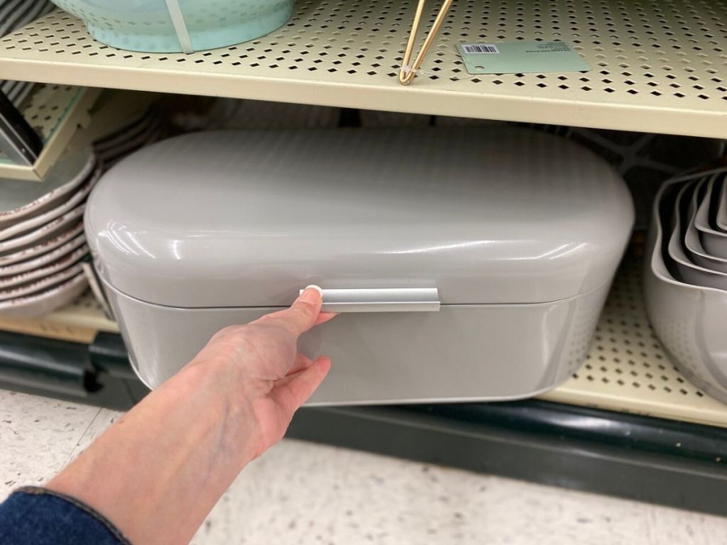 metal bread box on store shelf with human hand lifting it open partially 