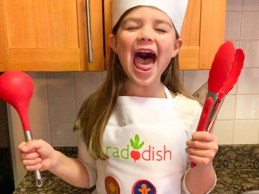 girl holding red ladle and tongs and wearing chef hat and raddish apron