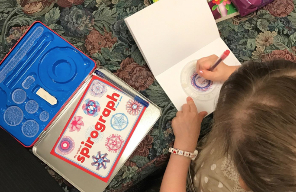 girl coloring on white paper with spirograph kit on table