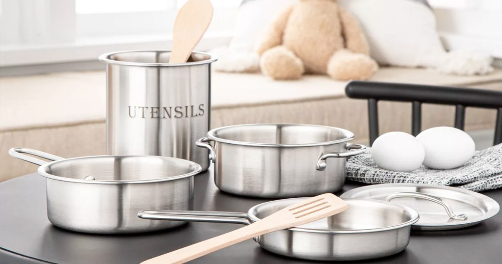 kids cookware set with pots, pans, lids, and wooden spoons on a black table in a playroom