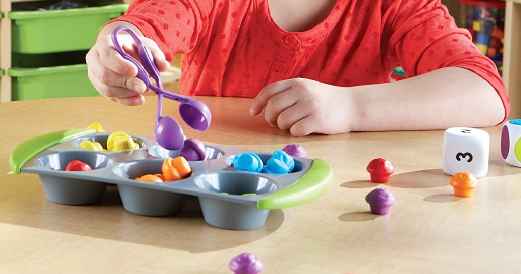 child using pair of purple plastic tongs to sort our small toys into cups of a muffin tin