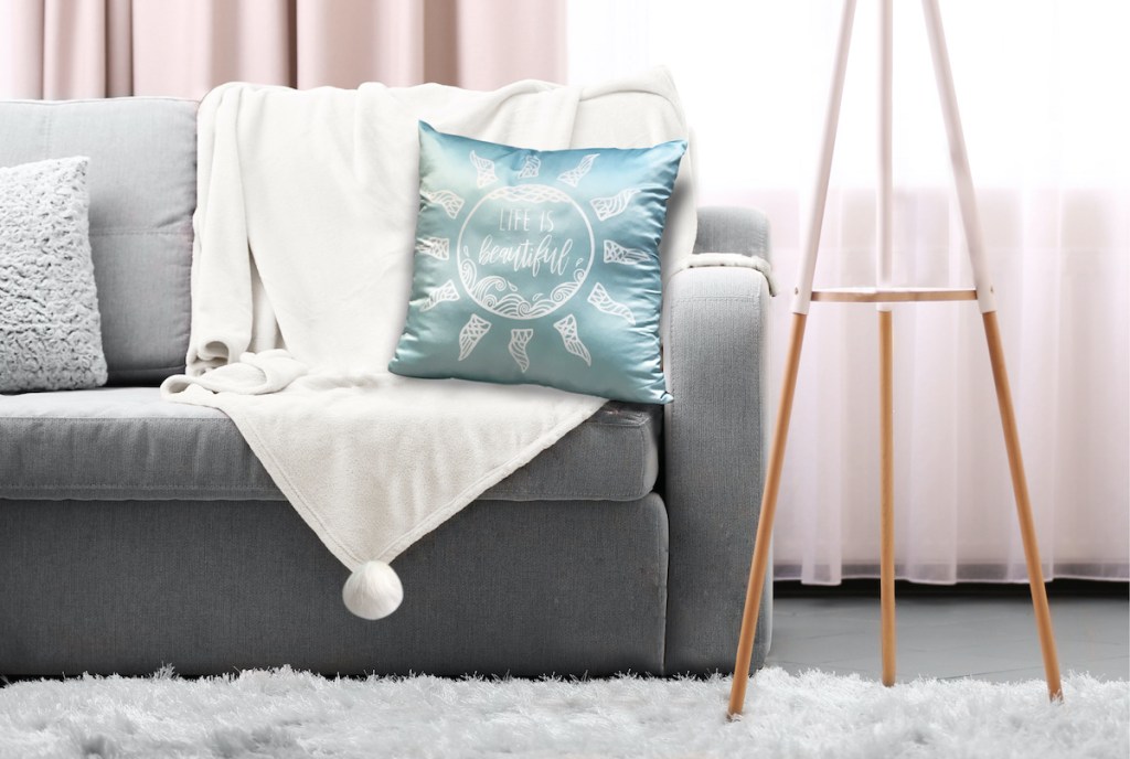 ice blue throw pillow with the saying "life is beautiful" on it with white throw blanket all on a gray couch in a living room