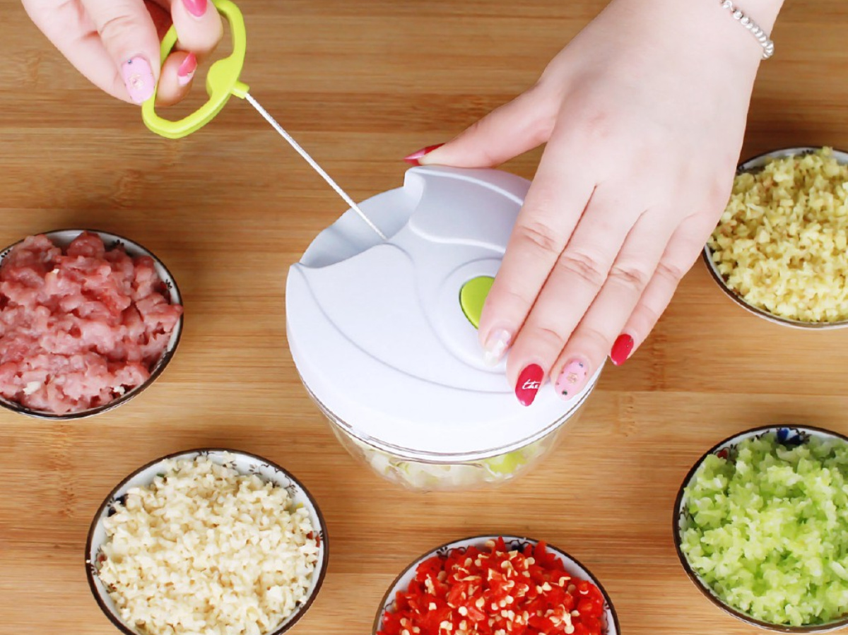 manicured hands holding white and green mini food chopper and pulling string with other hand, on top of wood cutting board, surrounded by bowls of chopped food