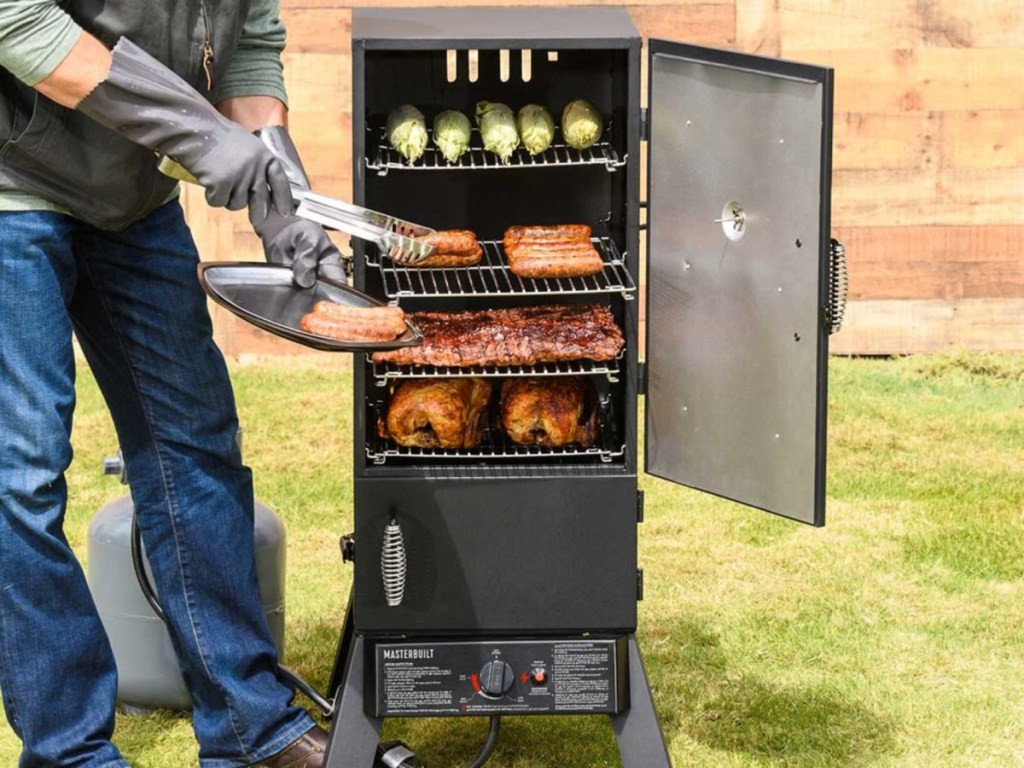 man wearing gloves holding a tray removing meat from a smoker with the door open