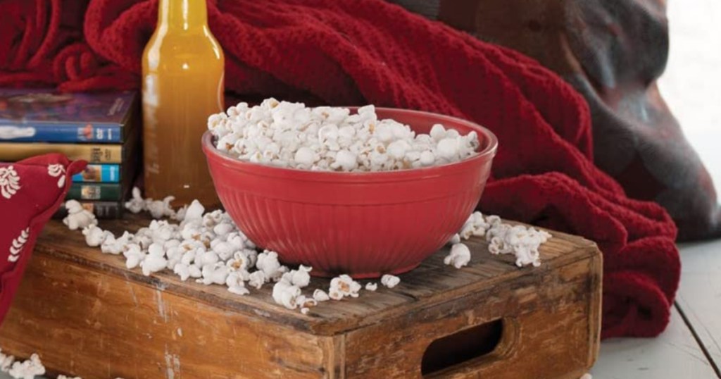 red bowl filled with popcorn on wooden table with orange soda and blankets surrounding it
