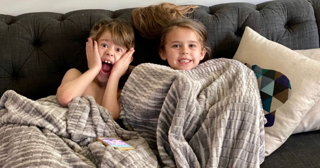 Two kids sitting on the couch under a blanket smiling