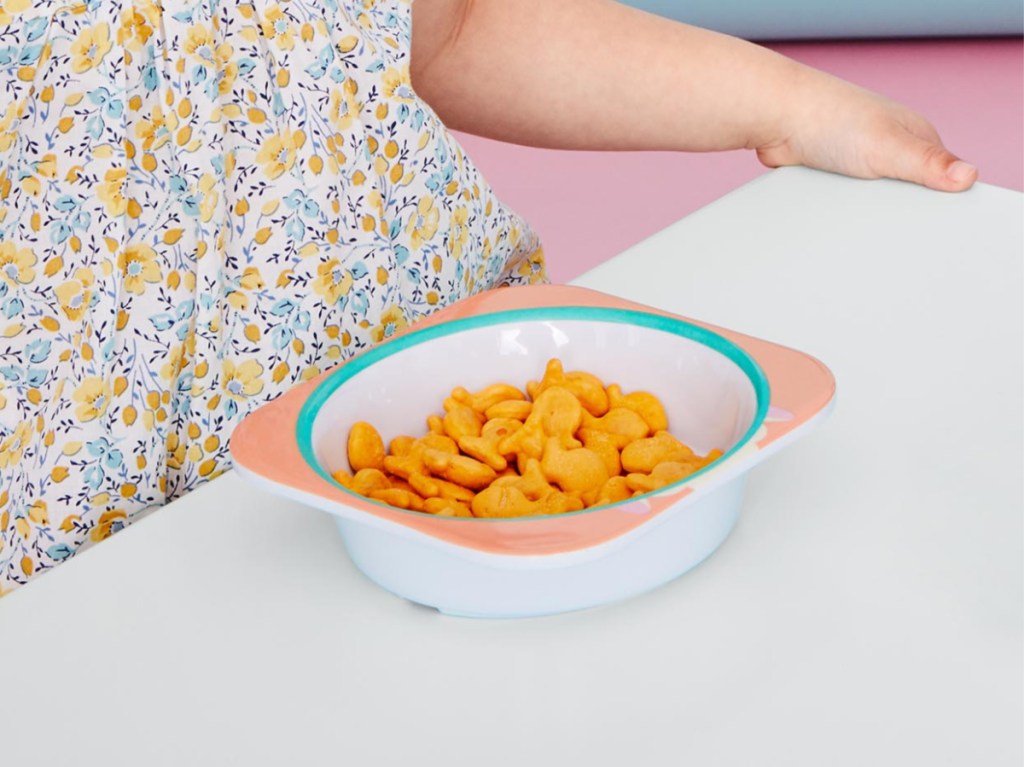 little girl in front of bowl of goldfish on white table