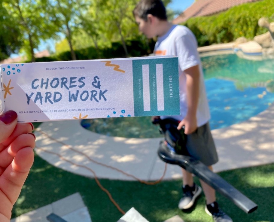 hand holding a chores and yard work coupon with kid holding leaf blower in background
