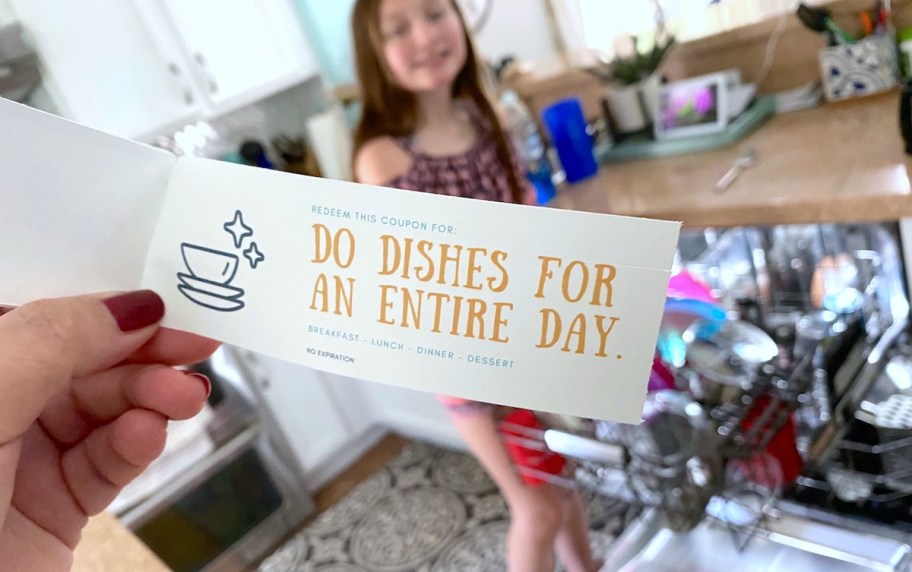 hand holding a diy voucher for doing the dishes and stading in front of dishwasher where a child is doing the favor for their mama