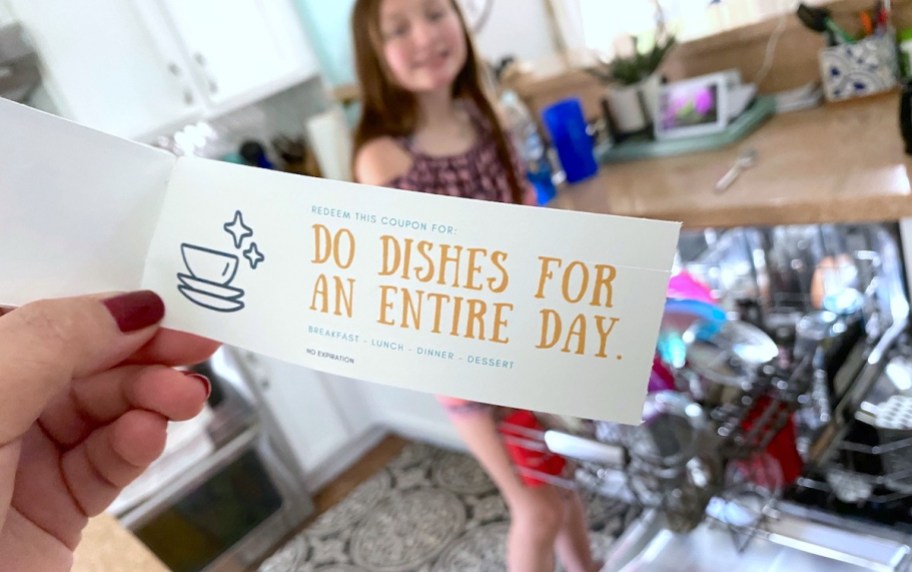 hand holding a diy voucher for doing the dishes and stading in front of dishwasher where a child is doing the favor for their mama