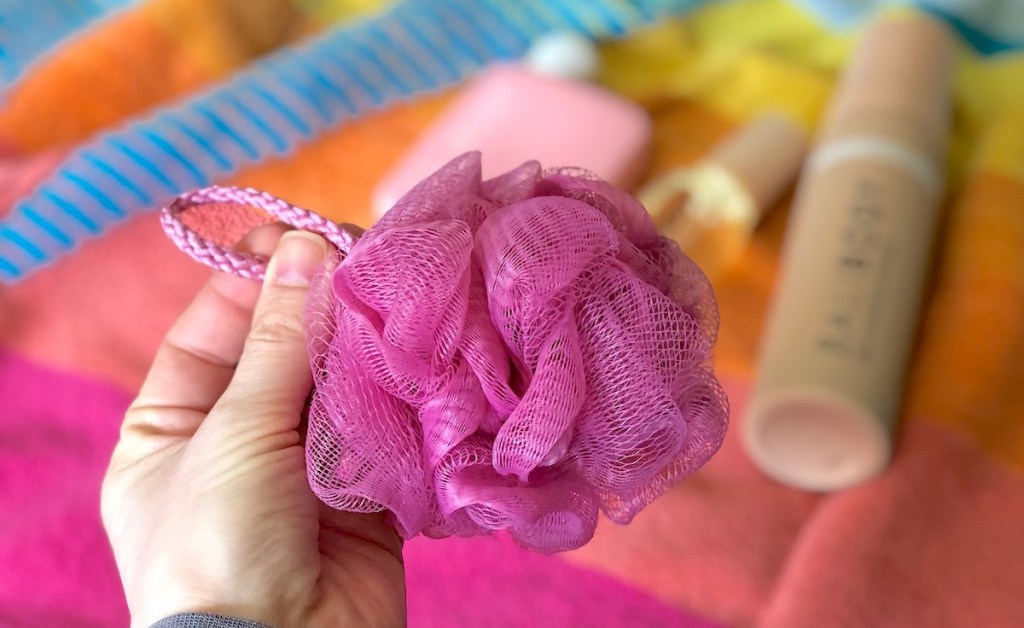 hand holding a bright pink loofa with lotions and bright colored towel in background