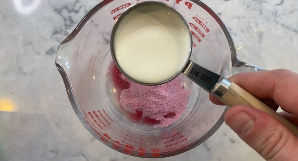 hand holding a measuring cup with milk over measuring cup with pink powder