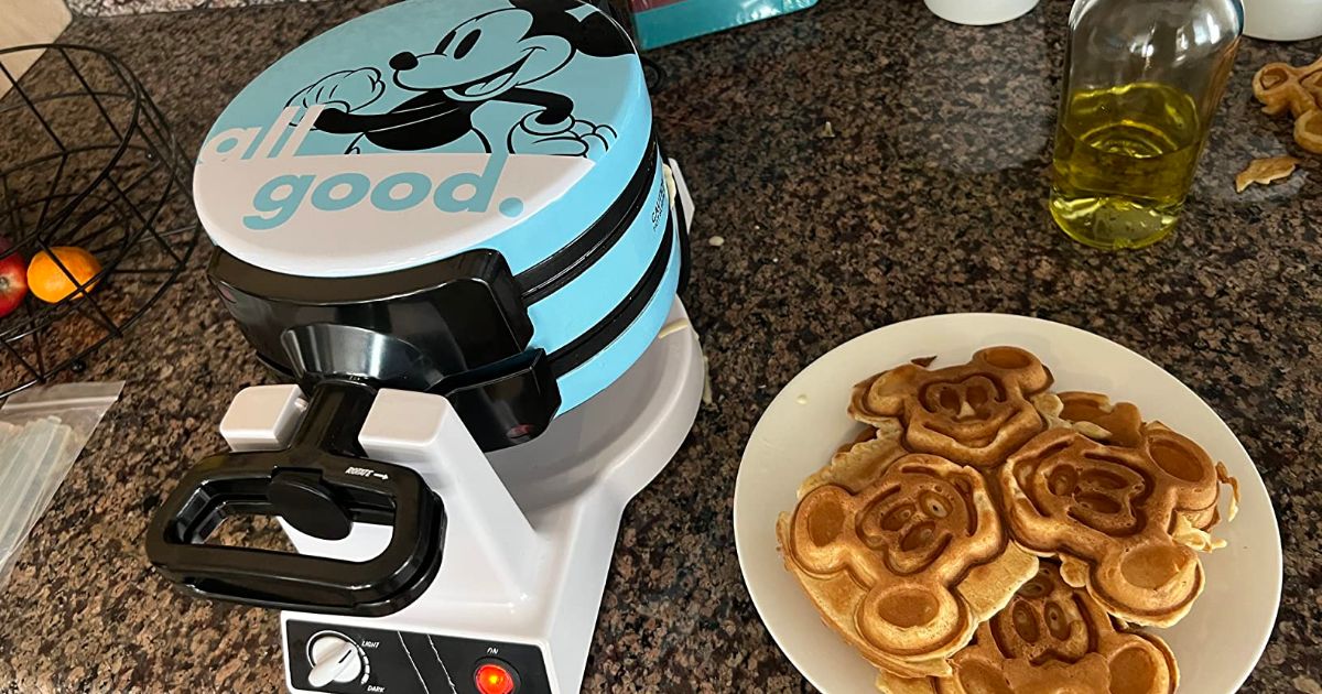 mickey & minnie flip waffle maker sitting on a kitchen counter next to a plate of golden delicios mickey-shaped waffles.