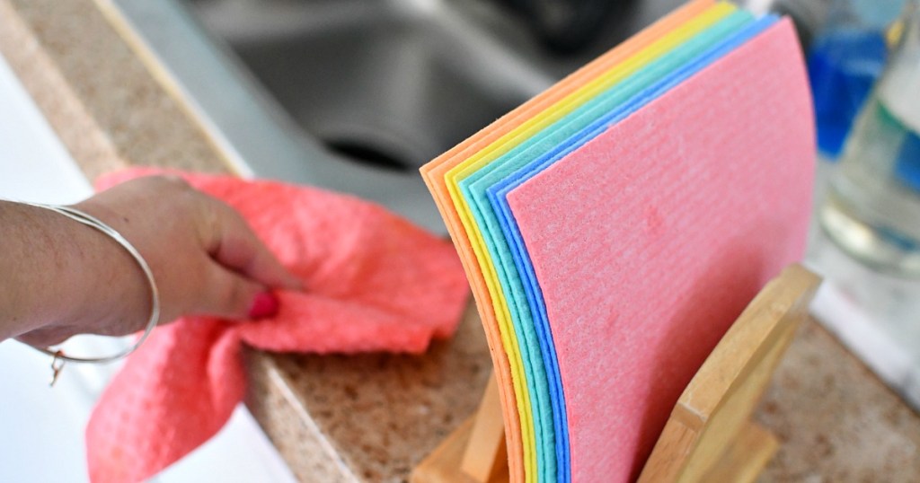 hand wiping down sink with pink swedish dishcloth and various colored ones in napkin holder