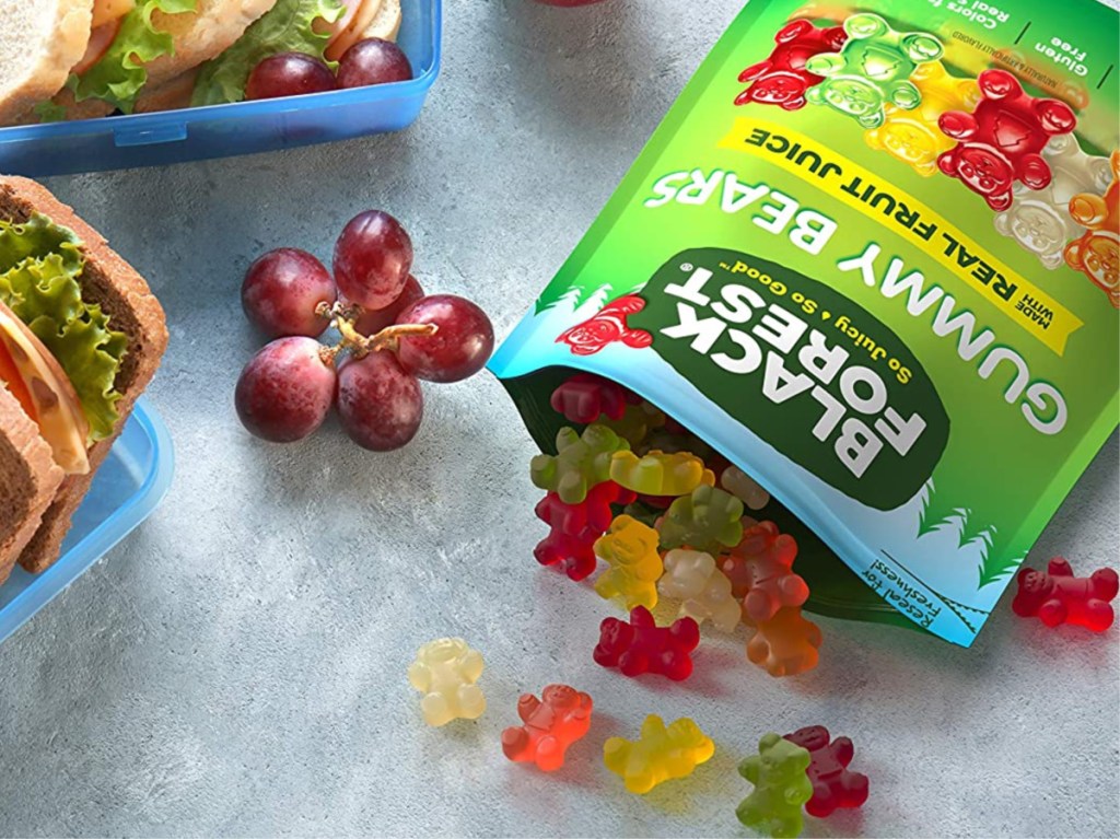 open bag of gummy bears with some fallen out onto counter, grapes, and sandwich in blue container