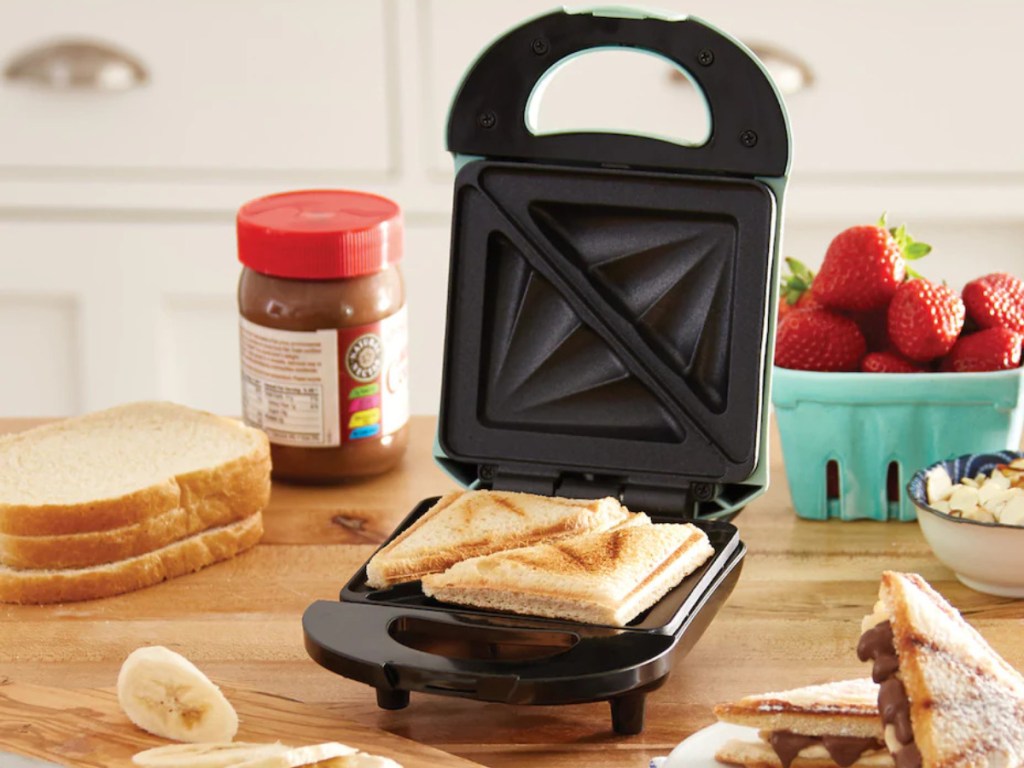 mini blue sandwich maker on wood counter with bread, sliced bananas, carton of strawberries, and chocolate spread