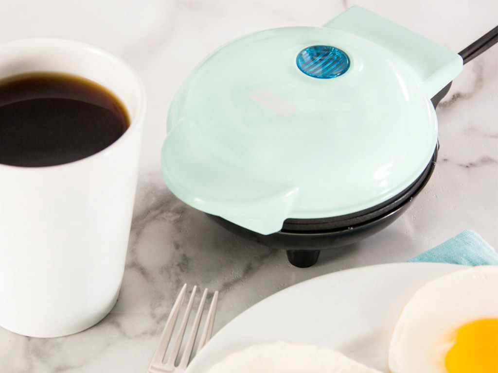 light blue mini griddle on table next to plate of eggs and white cup of coffee