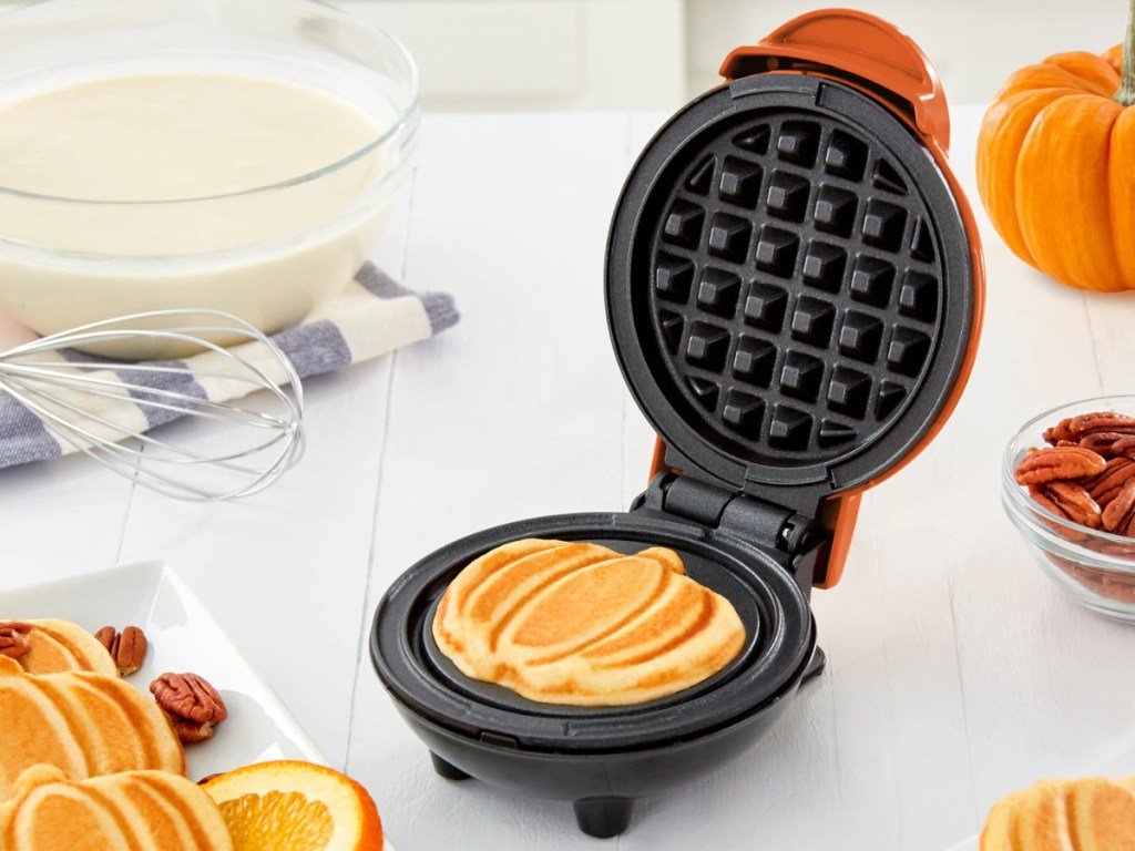 orange pumpkin-shaped waffle maker on white counter with pumpkin, bowl of batter, whisk, waffles