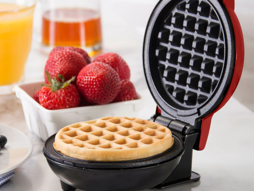red mini waffle maker with cooked waffle inside on counter with white bowl of strawberries and two glasses
