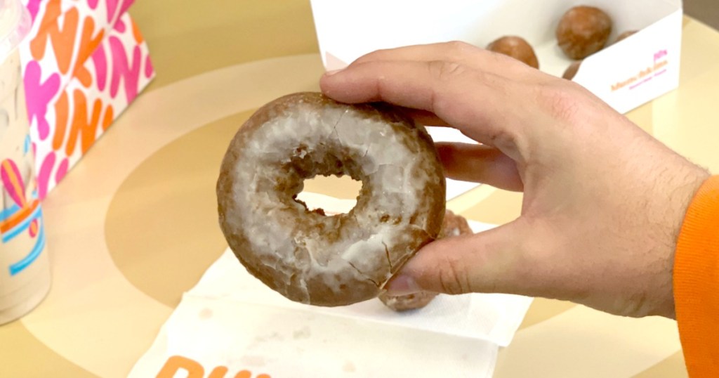Man's hand holding a classic style glazed donut near a box of donut holes