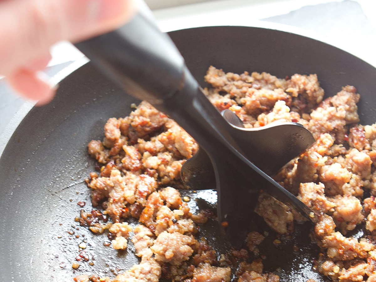 hand using black meat chopper on ground meat in black skillet pan
