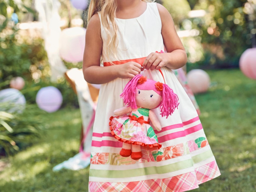 girl outside in yard holding doll wearing white and colorful striped dress with flower design