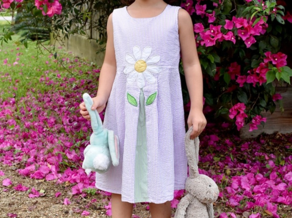 girl outside in front of flowers holding stuffed bunny animals in purple dress with large daisy on front