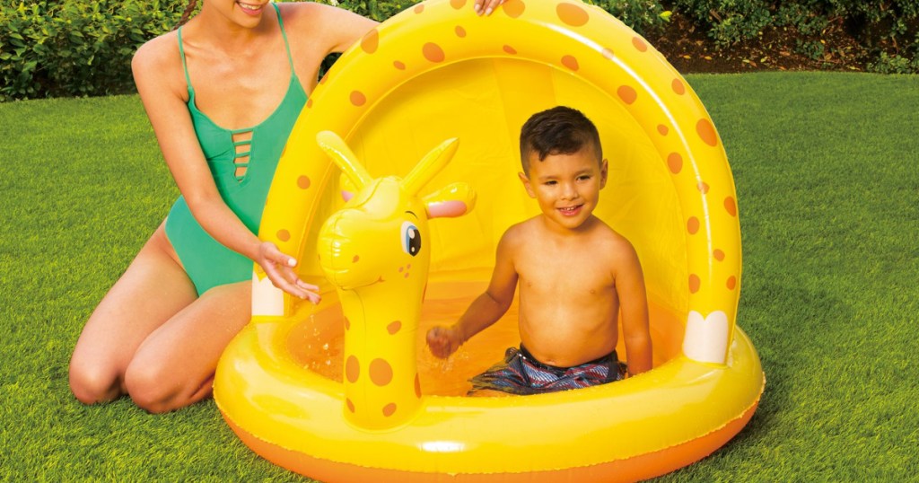 woman sitting on the grass next to a little boy in a giraffe pool