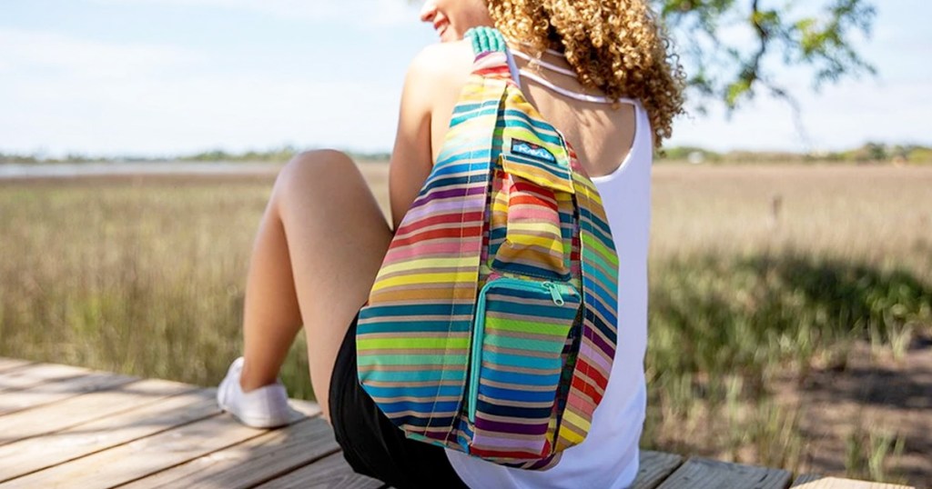 woman sitting on deck wearing a rainbow striped sling backpack overlooking a field