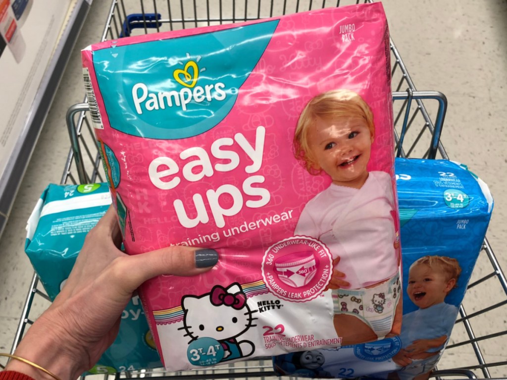 hand holding pink pack of girls training underwear above store cart full of diapers in store