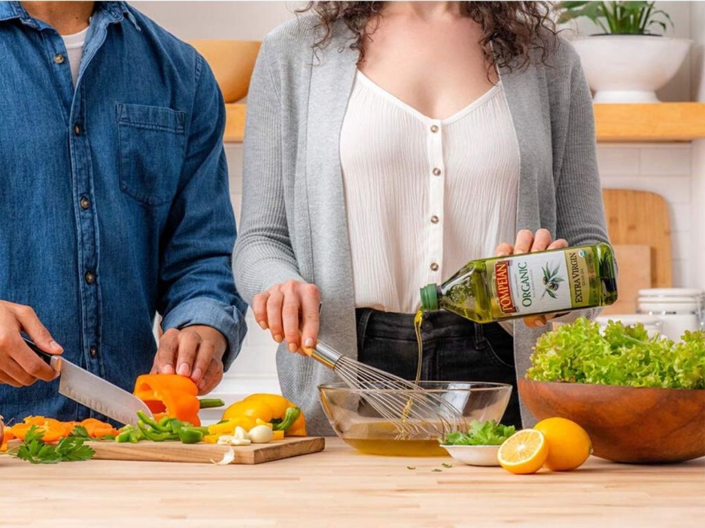 woman pouring bottle of olive oil into a bowl making salad dressing next to man chopping vegetables