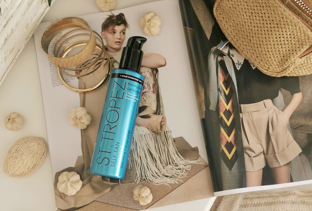 blue bottle of st tropez tanning mouse on top of open magazine with gold jewelry around it