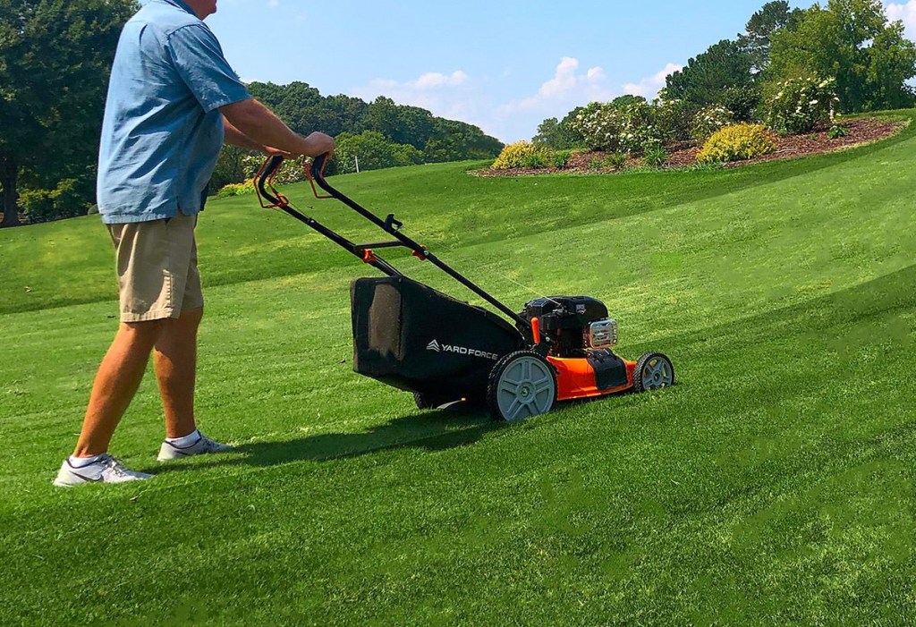 man pushing black and orange lawn mower up sloped hill of grass