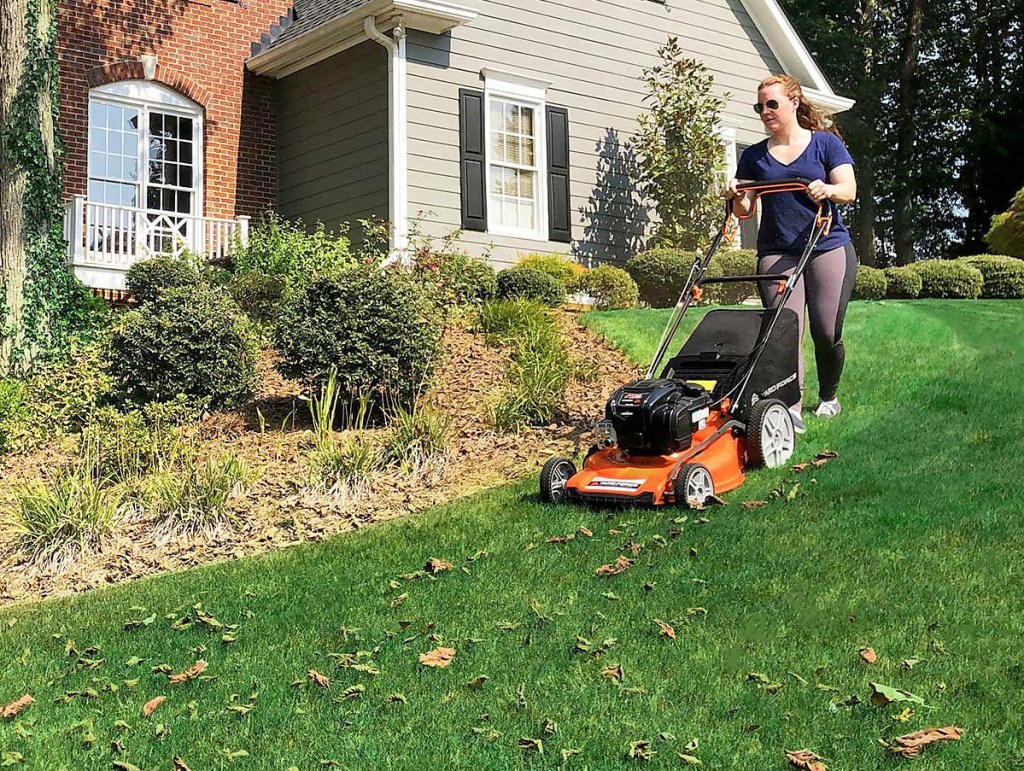 woman plushing orange and black lawn mower around garden in front of house