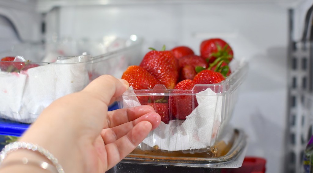 hand holding a clear container of fresh strawberries in fridge