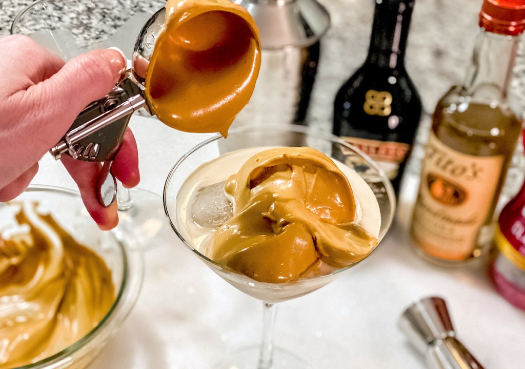 hand holding an ice cream scooper with brown whipped ingredients going into cocktail glass for dalgona martini